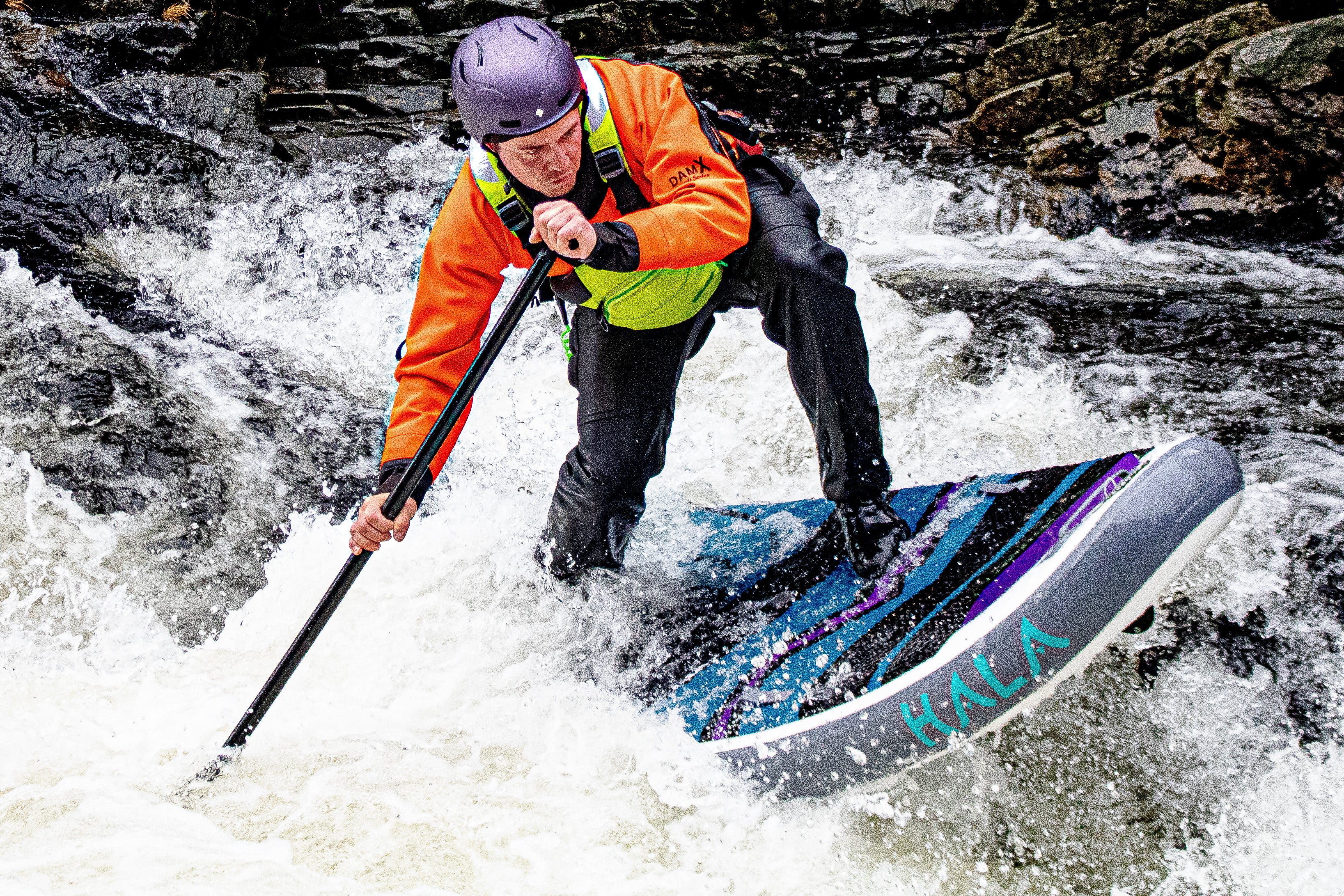 A man wearing a purple helmet, an orange dry top, and a neon green PFD (personal flotation device) is whitewater stand-up paddleboarding.