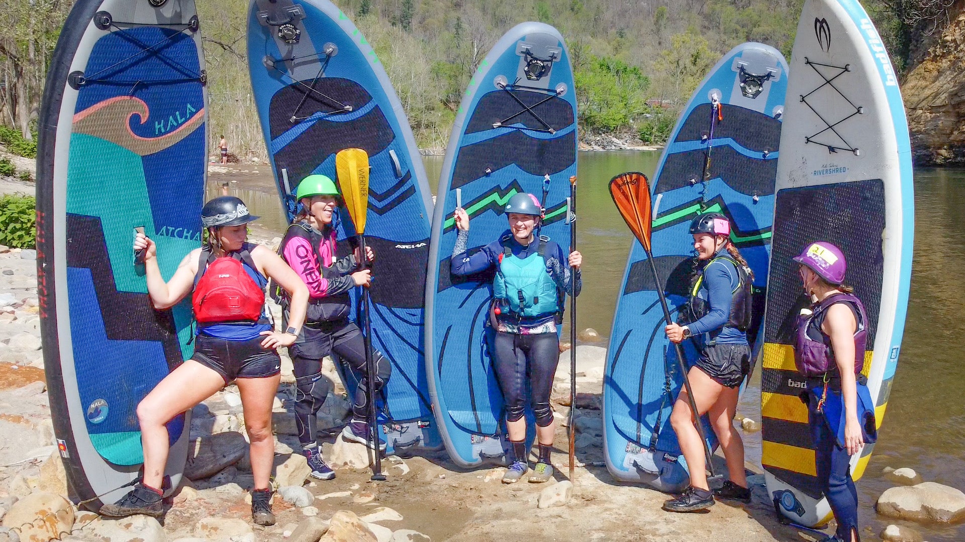 A group of five female paddleboarders in safety gear (helmets and PFDs) are standing next to their Hala stand-up paddleboards (SUPs) on a rocky riverbank.